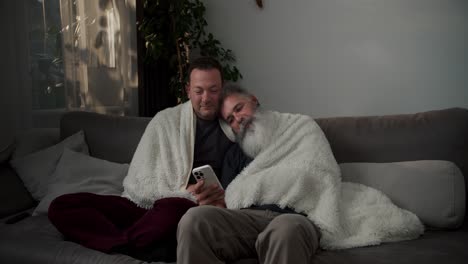 a happy brunette man sits on the sofa wrapped in a white woolen blanket with his elderly boyfriend with gray hair and a lush white beard they are looking at something on their white smartphone on a gray sofa in a modern apartment