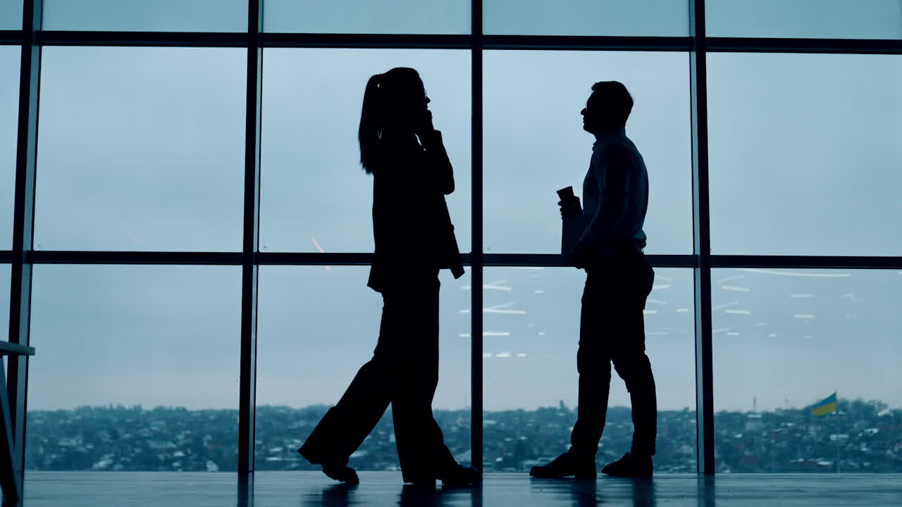 Male silhouette stands at the window drinking coffee. Female silhouette walks along speaking on the phone. Office employees at work.