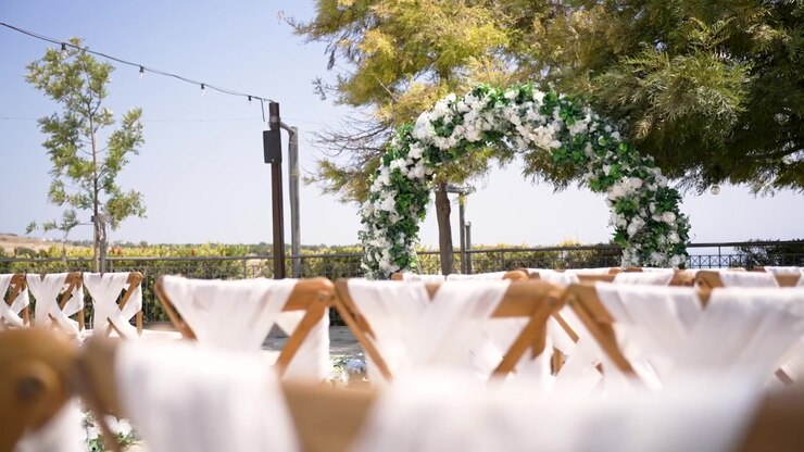 Et fantastisk udendørs bryllupssted med en blomsterbue og hvide siddepladser arrangeret til gæster. Omgivet af træer og et malerisk landskab er det perfekt til en romantisk ceremoni.
