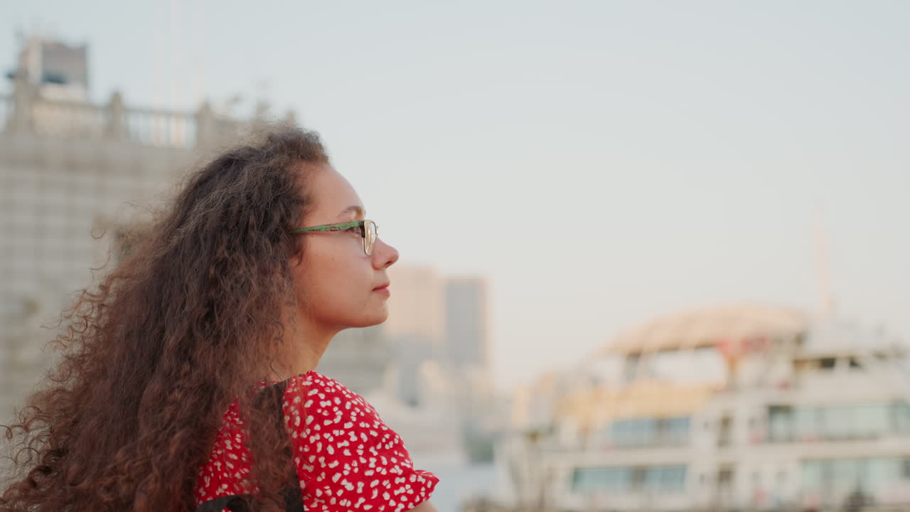 Donna in abito a pois che osserva le barche. Donna attenta in abito rosso che guarda navi lontane al tramonto. Signora contemplativa con occhiali e capelli ricci che si gode il paesaggio del porto cittadino al tramonto