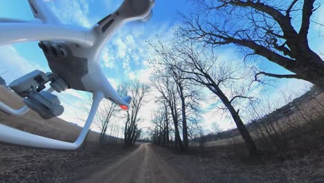 Close-up-Side-View-of-Dji-Phantom-4-Quadcopter-Drone-Flying-Forward-Over-A-Country-Gravel-Road-Among-Leafless-Trees