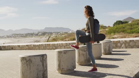 Frau-Mit-Gemischter-Rasse-Hält-Skateboard-In-Der-Hand-Und-Hält-An,-Um-Die-Aussicht-Am-Meer-Zu-Genießen