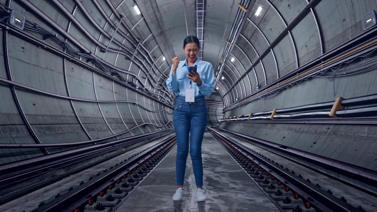 Full Body Of Asian Female With Her Smarphone In Underground Subway Tunnel, She Raises Her Fist Up With Screaming Goal