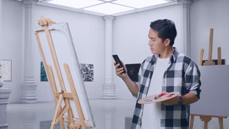 artista masculino asiático que encuentra inspiración en el teléfono inteligente mientras pinta en lienzo con pinturas al óleo y pincel en la galería