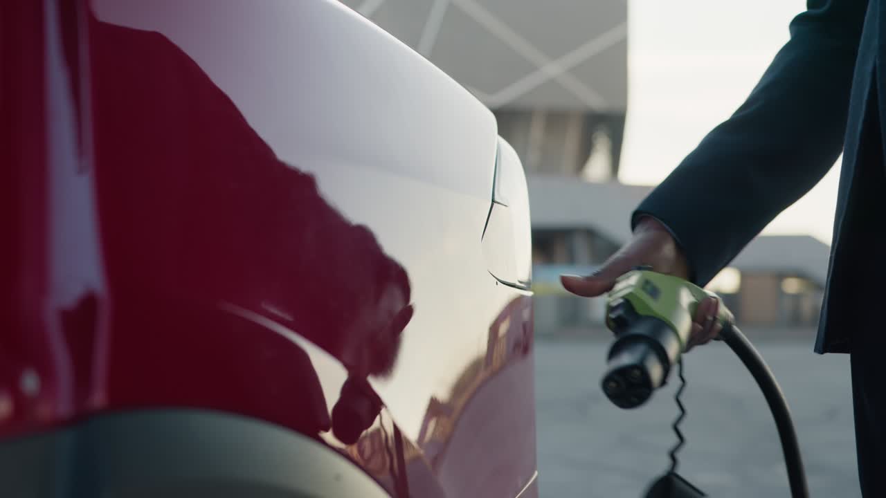 близкий взгляд на афроамериканца в формальном костюме, подключающего зарядный кабель к электрическому автомобилю. неузнаваемый бизнесмен, подключающий электрический автомобиль от зарядной станции. подключающий провод питания к автомобилю