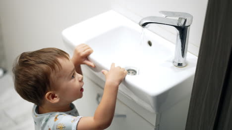 baby boy looks at water flowing from faucet. caucasian boy carefully touches the tap. top view.