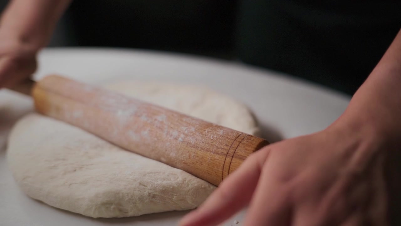 Hands roll dough with wooden roller on white surface. Flour dusts workspace as dough flattens. Process shows careful attention to detail. Ideal for cooking, bakery, or food preparation content