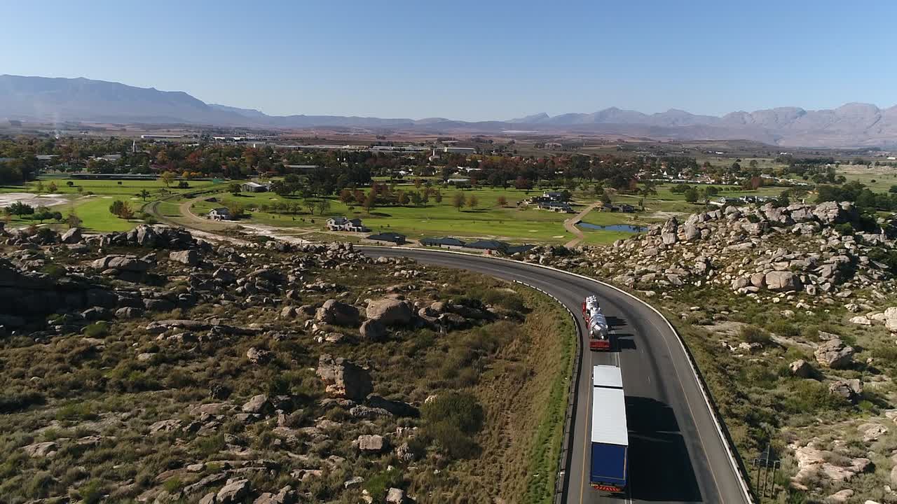 Vista aérea sigue camiones de transporte a través de terreno accidentado hacia un pequeño pueblo.