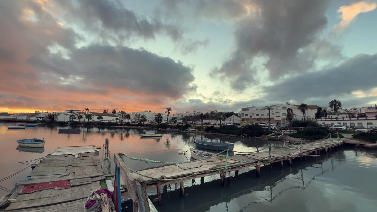 Wooden pier and several fishing boats reflecting on the calm water surface during a vibrant sunset in barbate, andalusia, spain, creating a picturesque and tranquil scene