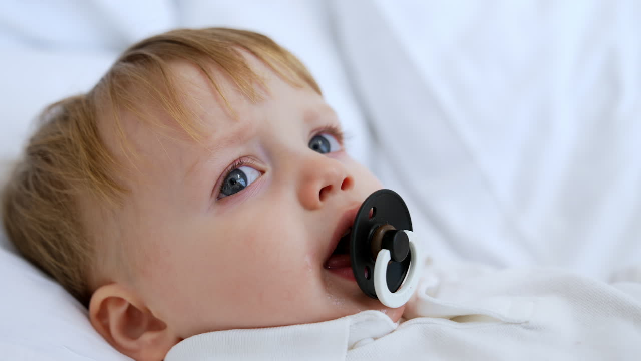 Cute Caucasian baby boy with fair hair lies on the pillow. Kid with pacifier is falling asleep. Close up.