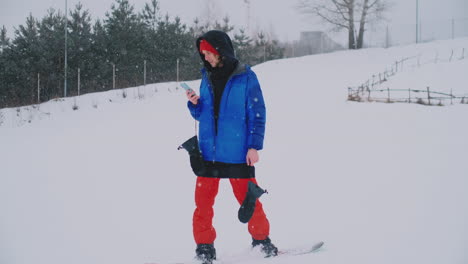 un snowboarder masculino monta la tabla en el esquí hasta la pista de nieve y escribe mensajes en el teléfono inteligente a sus amigos.