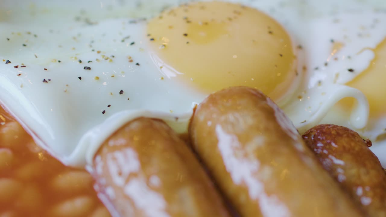 Knife and fork slice sausage beside eggs, bacon, and beans in bright, natural lighting