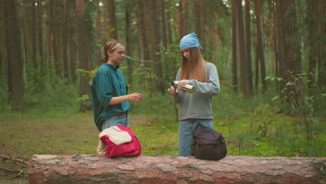 las hermanas disfrutan de una tranquila pausa al aire libre en un bosque exuberante, compartiendo una bebida caliente de un termo, una hermana vierte la bebida mientras la otra mira felizmente, rodeada de árboles y vegetación