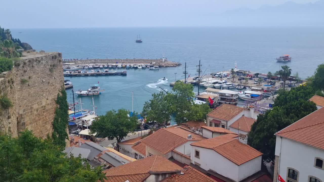 Scenic view over Antalya’s marina and the deep blue Mediterranean, with boats entering the port and a jet ski cruising by. In the foreground, the rooftops of the historic old town