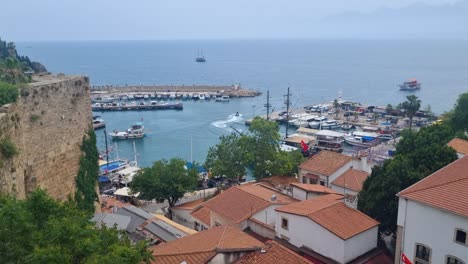 scenic view over antalya’s marina and the deep blue mediterranean, with boats entering the port and a jet ski cruising by. in the foreground, the rooftops of the historic old town