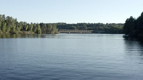 aerial view flying over calm lake towards pont de lantourne bridge in distance, forested shores, marcillac la croisille, france