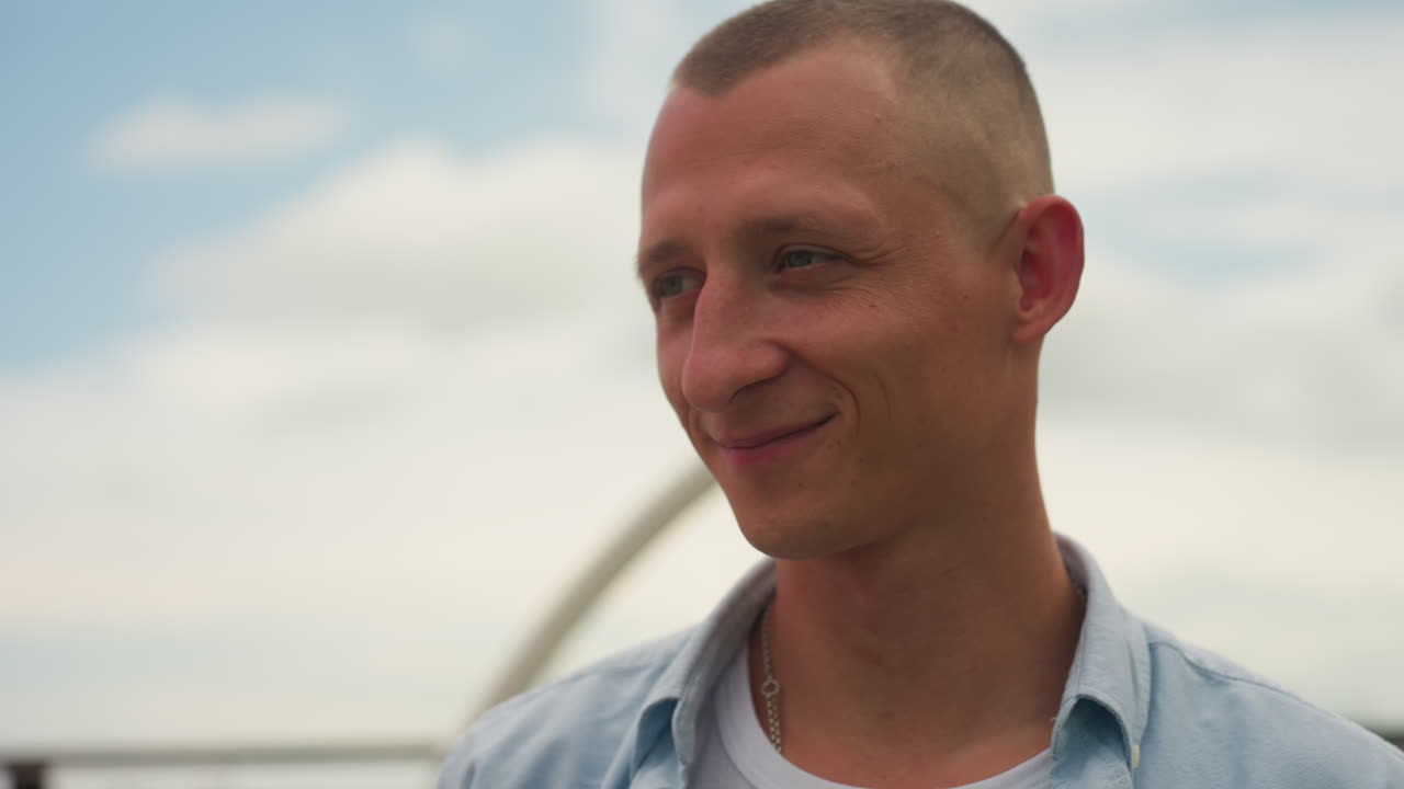 Uśmiechnięty mężczyzna na dachu pod gołym niebem, w jasnej koszuli dżinsowej i z krótko ostrzyżonymi włosami, miękkie światło słoneczne z oddalonym dźwigiem i miejską panoramą, szczera, zrelaksowana postawa i delikatny uśmiech wyrażający spokojną satysfakcję