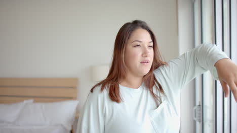 Thoughtful-plus-size-biracial-woman-looking-out-of-bedroom-window-in-the-morning,-slow-motion