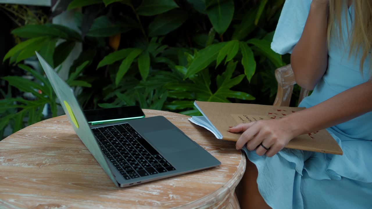 파란 드레스를 입은 소녀가 노트북 뒤에서 온라인에서 원격으로 작업하고 초록색 식물이 있는 뒷마당을 스크린으로 보고 있습니다.