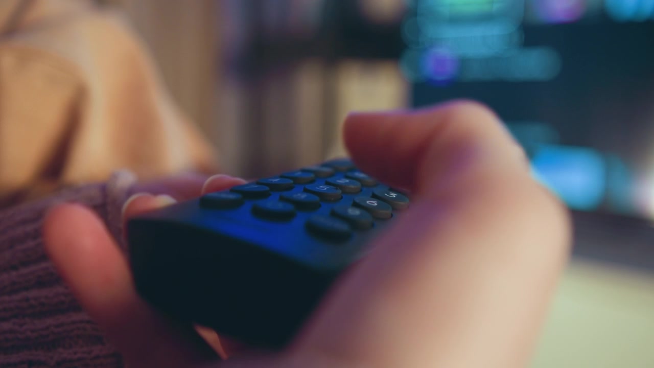 Close-up hands gripping remote control with thumb pressing buttons, slight motion blur and depressed keys suggesting active channel or volume change, set in cozy living room with warm evening light