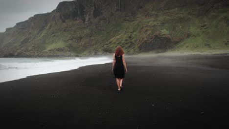 Schöne-Rothaarige-Frau-Im-Schwarzen-Kleid,-Die-Am-Schwarzen-Sandstrand-In-Island-Spaziert,-Kamerafahrt