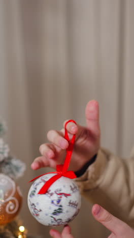 teenage boy holds in his hands a beautiful ball toy for christmas tree. kid decorating the fir tree with toy and garlands indoors. vertical video