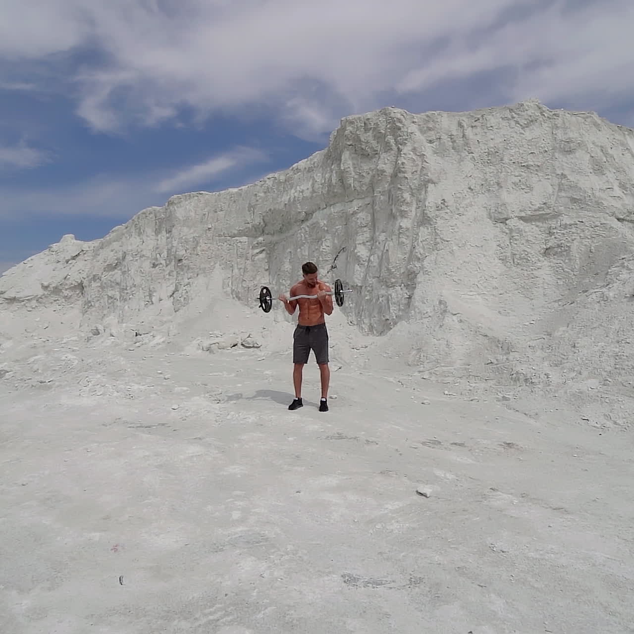 Sporty man lifting heavy barbell in the mountains. Bodybuilder doing his workout on the white background of a rocky hill. Slow motion.