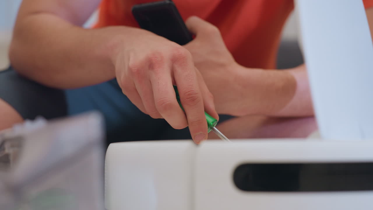 Close up of vacuum repairer in orange shirt seated on floor holding phone while inspecting robot vacuum cleaner with open lid showing repair process and attention to technical details