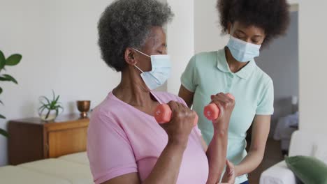 african american female physiotherapist wearing face mask helping senior female patient exercise