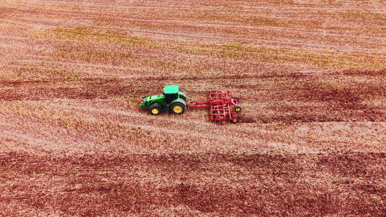 Macchina verde con erpice rosso che si muove attraverso un campo di stoppie rosso-marrone, tracce di trattore visibili nel terreno polveroso sotto una luce diffusa. Vista laterale di un trattore agricolo verde che ara un campo secco.