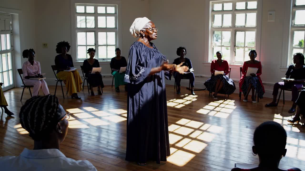 A woman speaks passionately in a sunlit room, surrounded by an attentive group