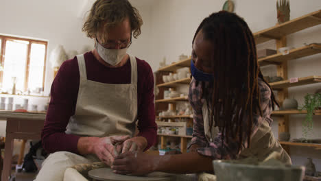 diverse male and female potters wearing face masks creating pottery on potters wheel at pottery stud