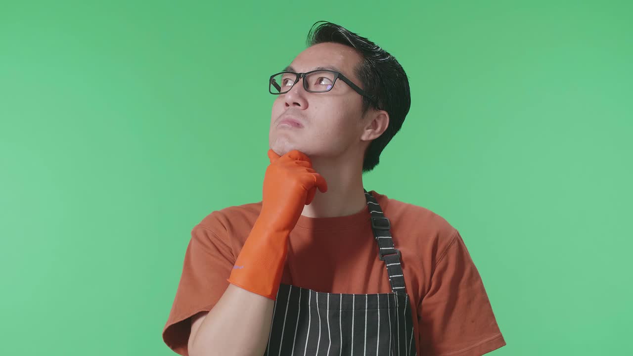 Close Up Of Asian Male Housekeeper With An Apron Thinking About Something And Looking Around While Standing In The Green Screen Studio