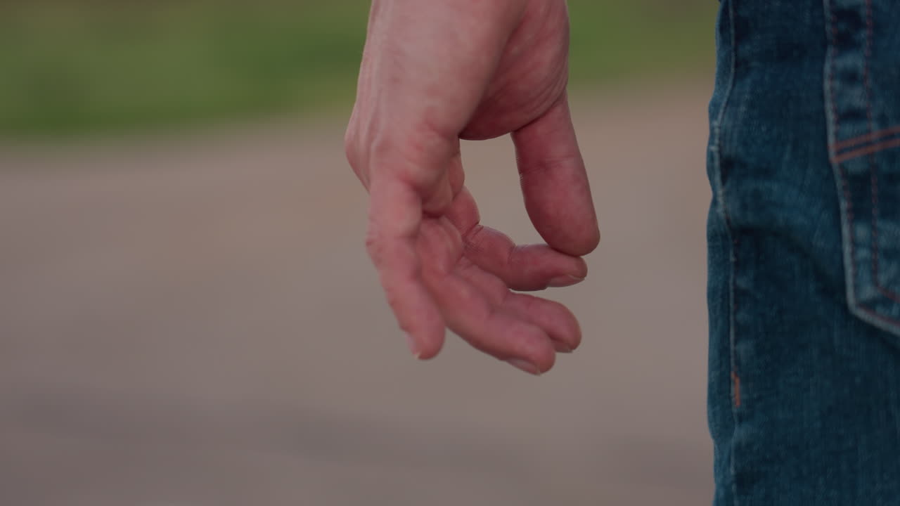 side view of man wearing pale shirt and denim jeans with shoulder bag gesturing with hand by roadside near parked car and green field bordered by distant trees on rural road under soft evening light
