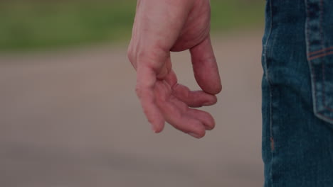 side view of man wearing pale shirt and denim jeans with shoulder bag gesturing with hand by roadside near parked car and green field bordered by distant trees on rural road under soft evening light