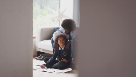 Pre-teen-boy-sitting-on-the-floor-at-home-doing-homework-with-his-father-sitting-on-the-sofa-behind-him-helping,-looking-at-each-other,-seen-from-doorway