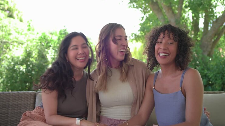 Diverse amiche sedute sul divano in patio, rivolte verso la fotocamera, che ridono insieme tenendo in mano degli orologi.