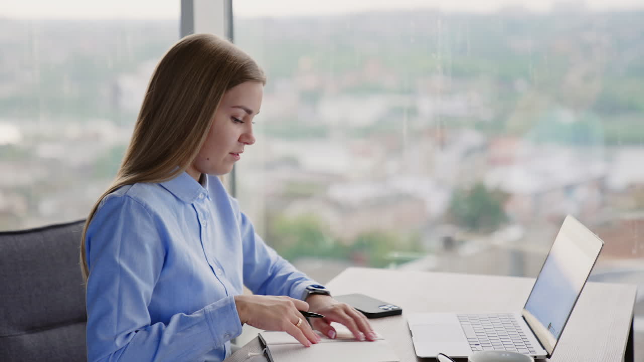 Female office employee sits at the table with laptop. Woman looks into her paper notebook smiling. Blurred backdrop.