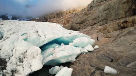 Photographer-taking-photos-of-spectacular-ice-cave,-rugged-terrain-ice-and-rock,-New-Zealand-Alps---drone-shot