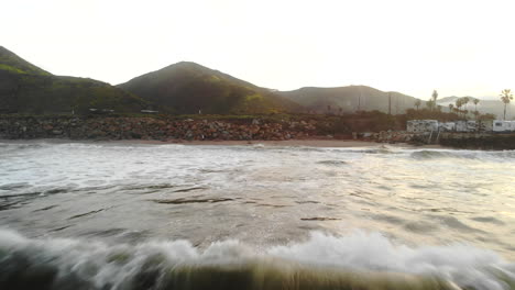 Aerial-drone-flying-low-over-the-pacific-ocean-waves-on-the-beach-at-sunrise-with-palm-trees-and-green-mountains-in-the-background-in-Ventura,-California