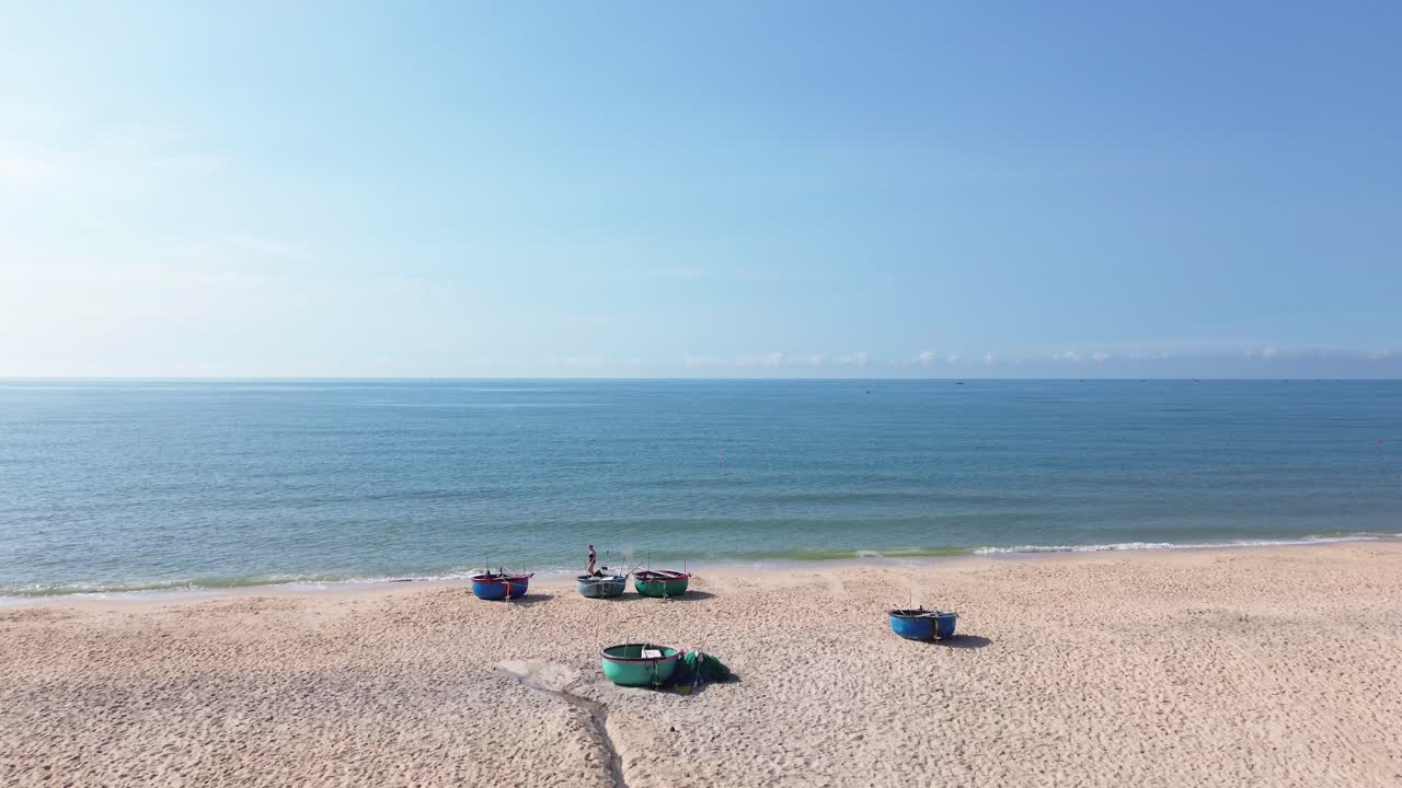 Drone moves from Mui Ne beach toward the sea, flying low over parked bucket boats with a person walking nearby along the shoreline.