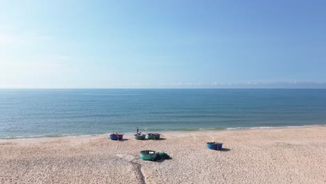 drone moves from mui ne beach toward the sea, flying low over parked bucket boats with a person walking nearby along the shoreline.