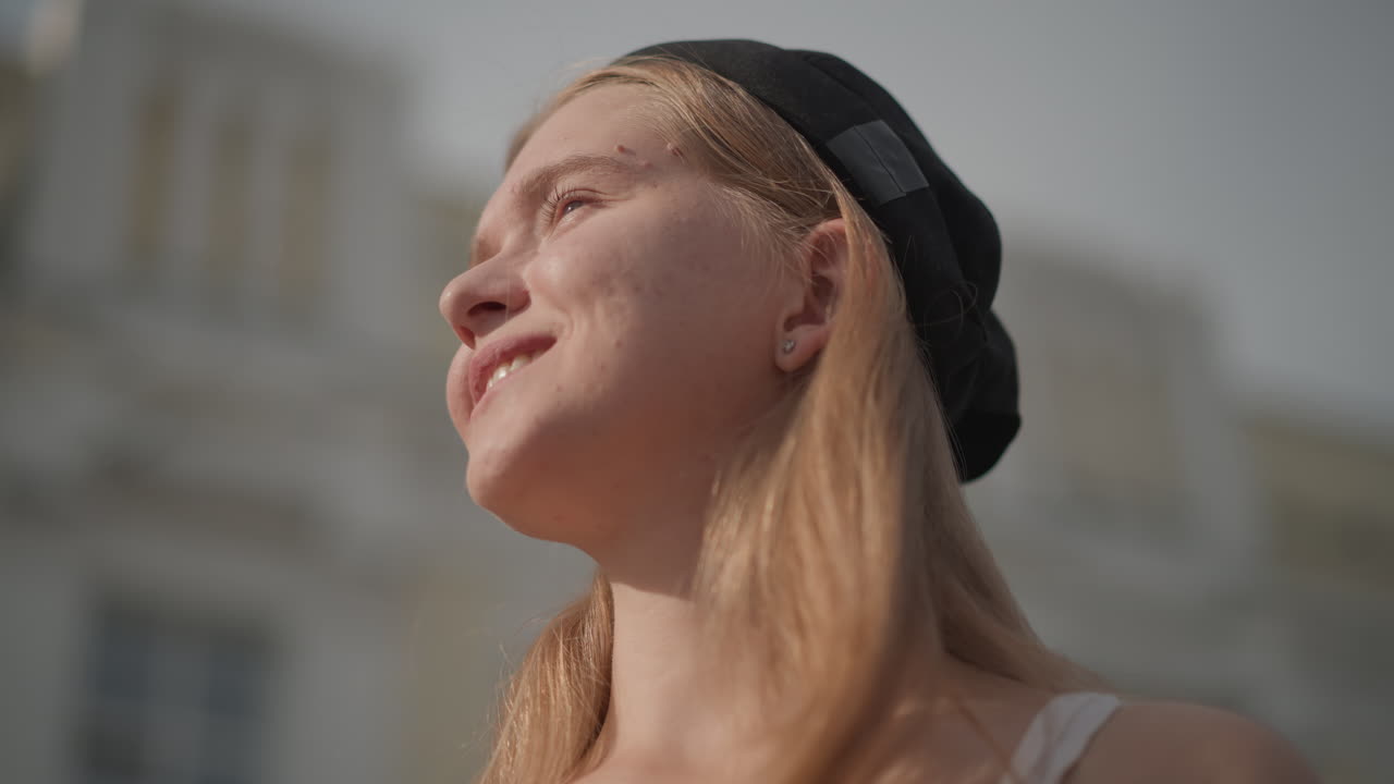 Solrik gateportrett av en smilende, hvit tenåringsdanser med hendene bak hodet, fregner som fanger varmt lys, liten lue, blikket oppover mot blå himmel, myke urbane bygninger uskarpe i bakgrunnen, oppriktig glede