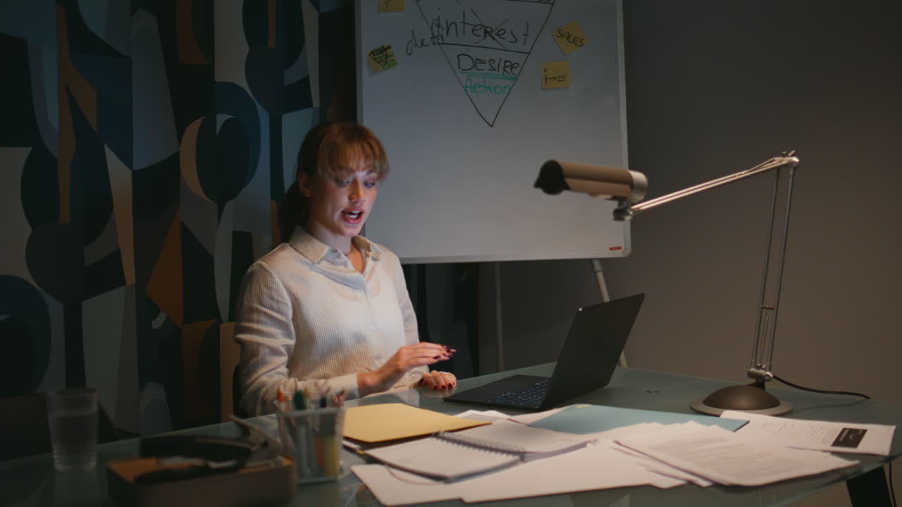 Primo piano di una signora che saluta tramite videochiamata serale utilizzando un laptop in un ufficio buio. Donna che parla