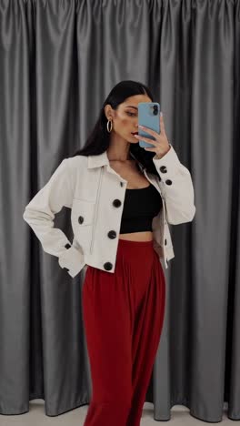 una mujer joven se toma fotos con su teléfono inteligente en un camerino, con una chaqueta blanca y pantalones rojos, haciendo varias poses para mostrar la ropa