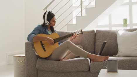 video of happy biracial woman in headphones sitting on sofa and playing guitar