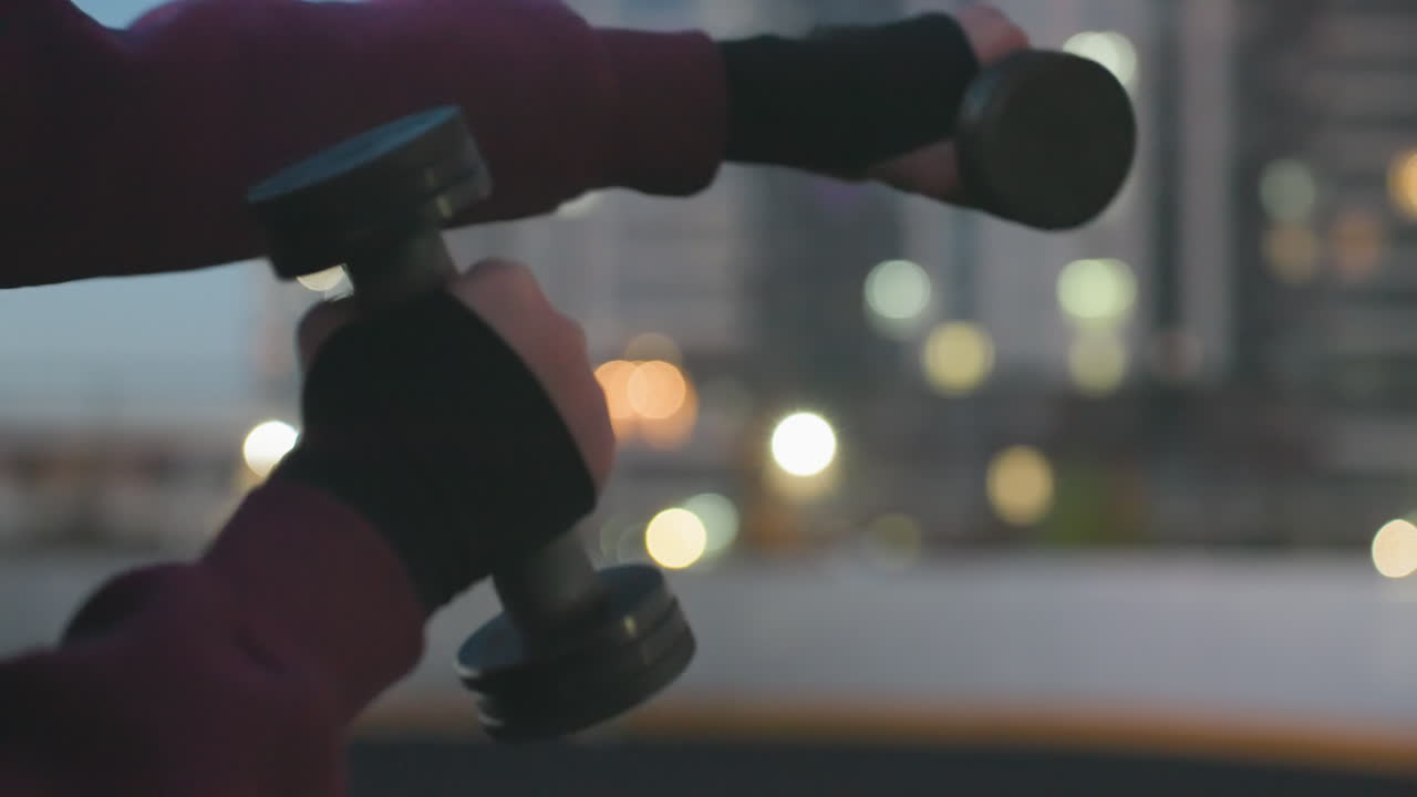 Hand view of fitness enthusiast boxing dumbbells punching air on black asphalt sports court beside white barrier fence at dusk against urban high rise backdrop wearing maroon hoodie white sneakers