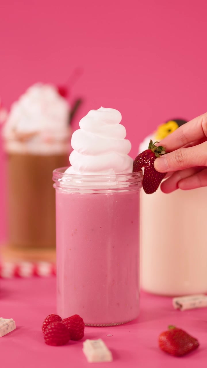 Una mano posiziona una fragola su un milkshake rosa guarnito con panna montata, bevande dessert colorate, illuminazione brillante.