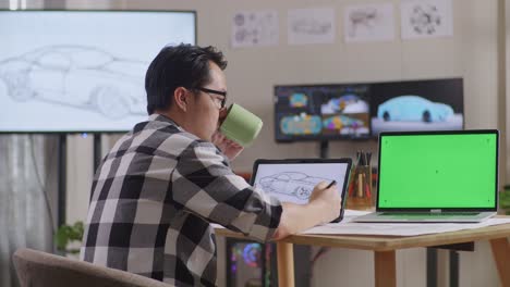 아시아 남성이 커피를 마시며 테이블릿에 자동차 컨셉을 그리는 측면 뷰, tv와 컴퓨터를 가진 스튜디오에서 녹색 스크린 노트북 에 3d 전기 자동차 모델을 표시합니다.