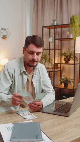 rich happy caucasian man counting money cash using laptop calculate domestic bills at home table